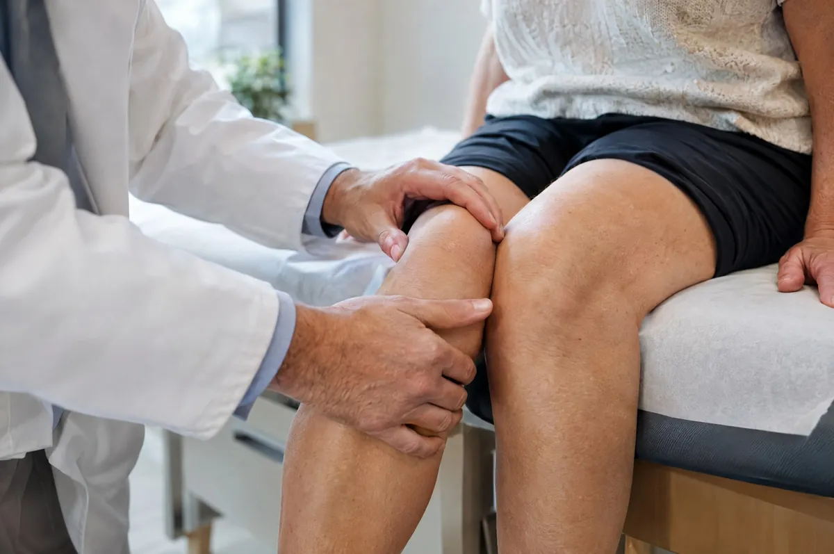 Symbolbild: Ärztliche Hände untersuchen das Knie einer sitzenden Person
